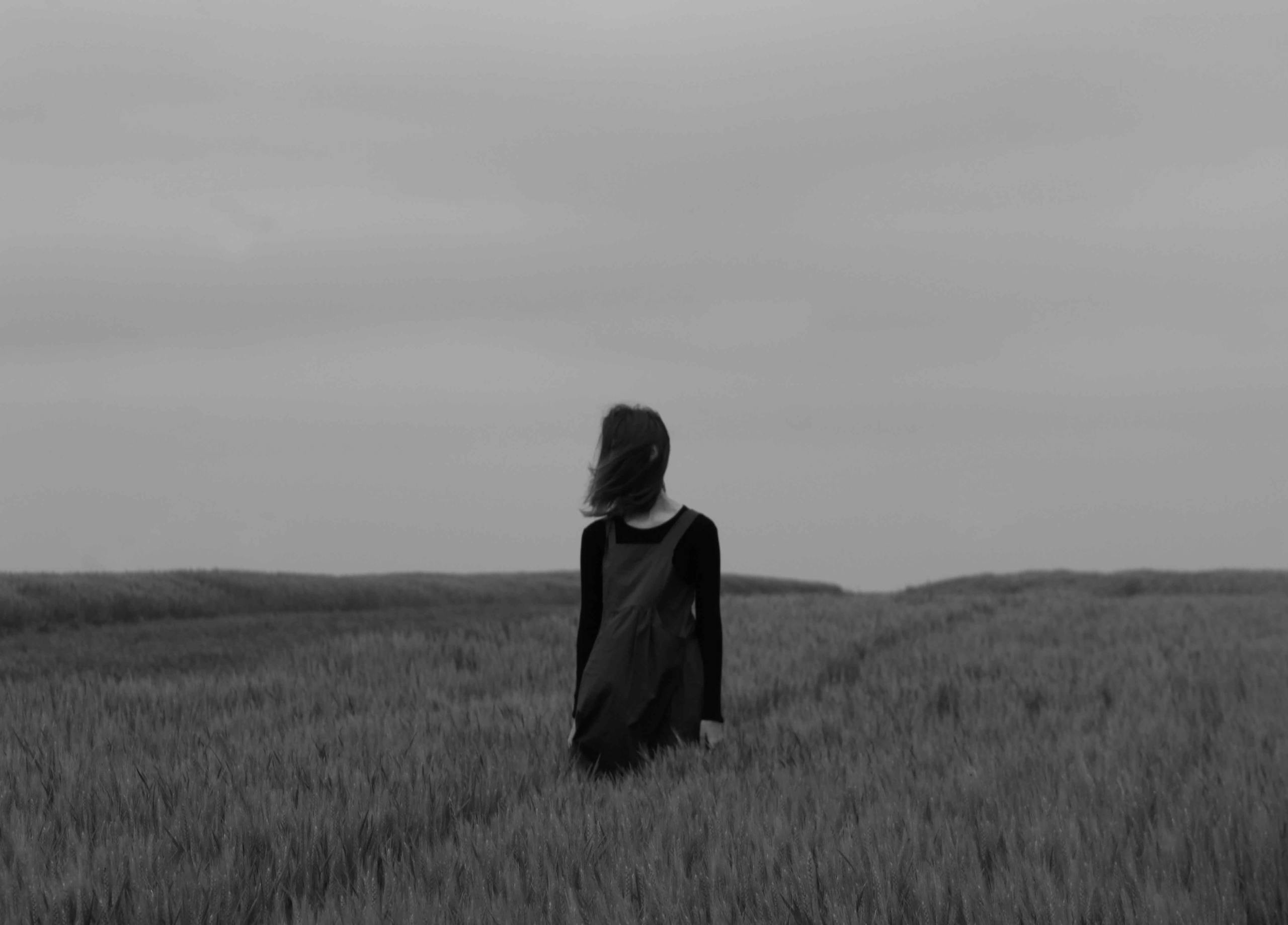 Black and white image of a woman standing alone in a vast wheat field under a cloudy sky.
